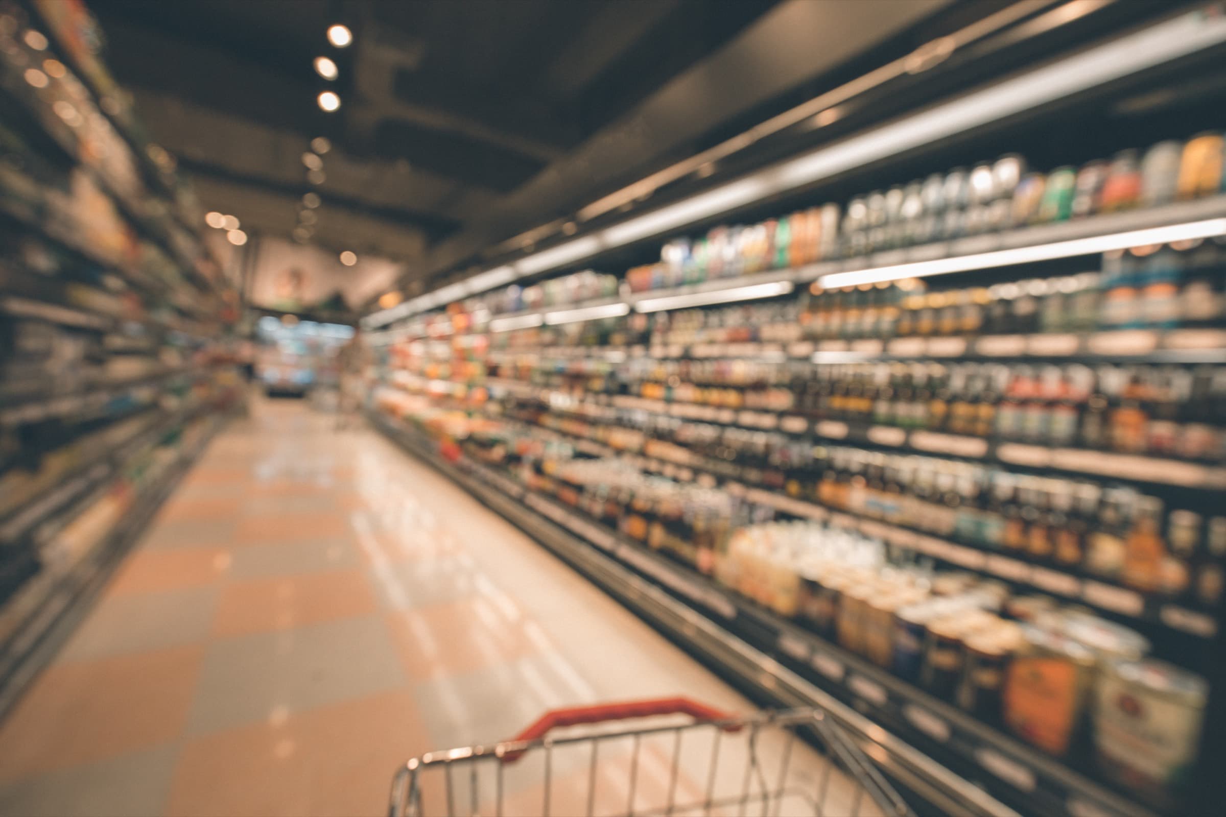 A supermarket aisle seen from behind a shopping trolley, shelves of packaged goods running away down the right.